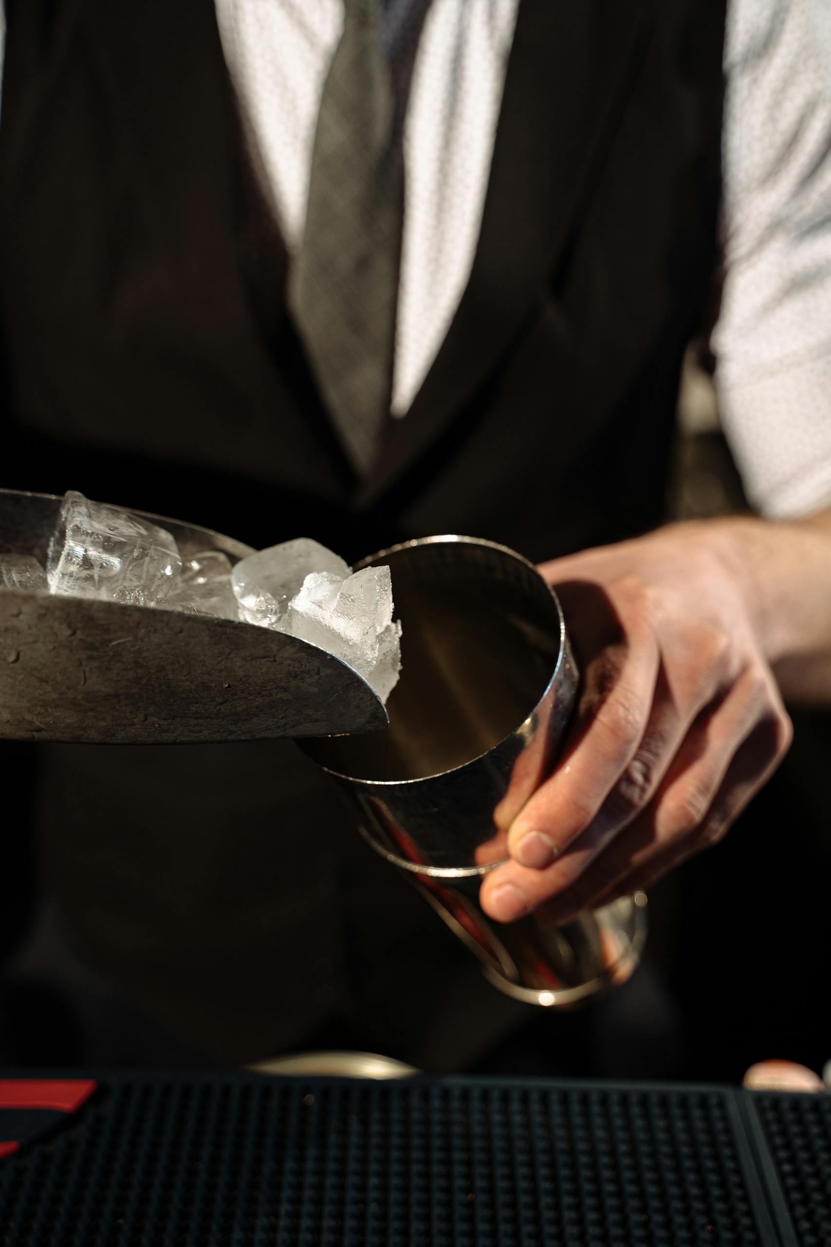 Bartender adds ice into a shaker using a scoop in a dimly lit bar setting.